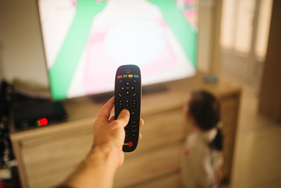 Copil urmărește canale tv romanesti la televizor în timp ce un adult ține telecomanda